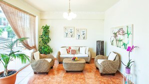 A living room with wicker furniture, a sofa, and a coffee table with fruit. There are potted plants and artwork on the walls. - Casa Di Mare Poseidon (Katerini)