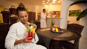 A person in a white robe holding a drink with fruit garnish, seated at a table with a fruit bowl. - Wellness Parc Hotel Ruipacherhof (Tirolo)