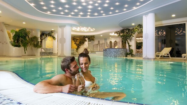 A couple in a large indoor swimming pool with a tiled floor, surrounded by columns and plants. - Wellness Parc Hotel Ruipacherhof (Tirolo)