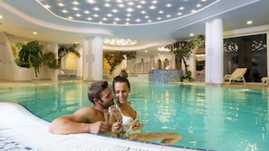 Una pareja en una amplia piscina interior con suelo de baldosas, rodeada de columnas y plantas.