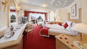 A hotel room with a large bed, a desk with a chair, a small table, and a large mirror. There is a window with a view of trees and mountains. - Wellness Parc Hotel Ruipacherhof (Tirolo)
