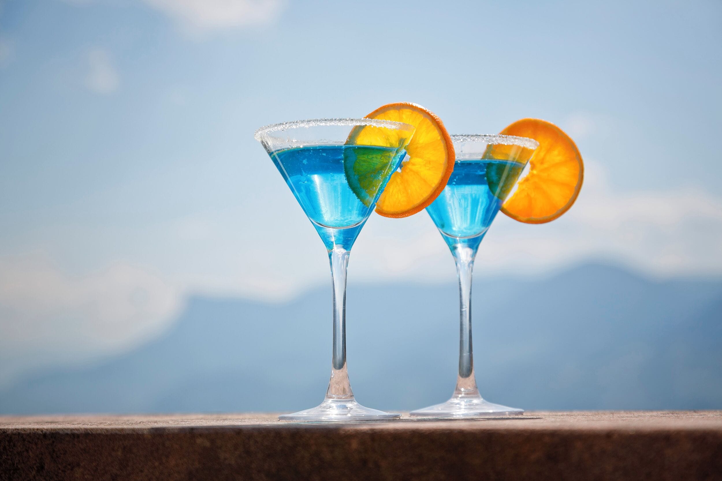 Deux verres à martini contenant un liquide bleu et des tranches d’orange, posés sur une étagère avec une chaîne de montagnes en arrière-plan.