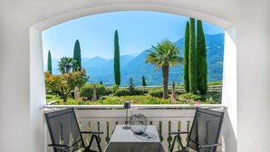 Varanda com mesa e duas cadeiras, com vista para um jardim ajardinado, árvores altas e montanhas.