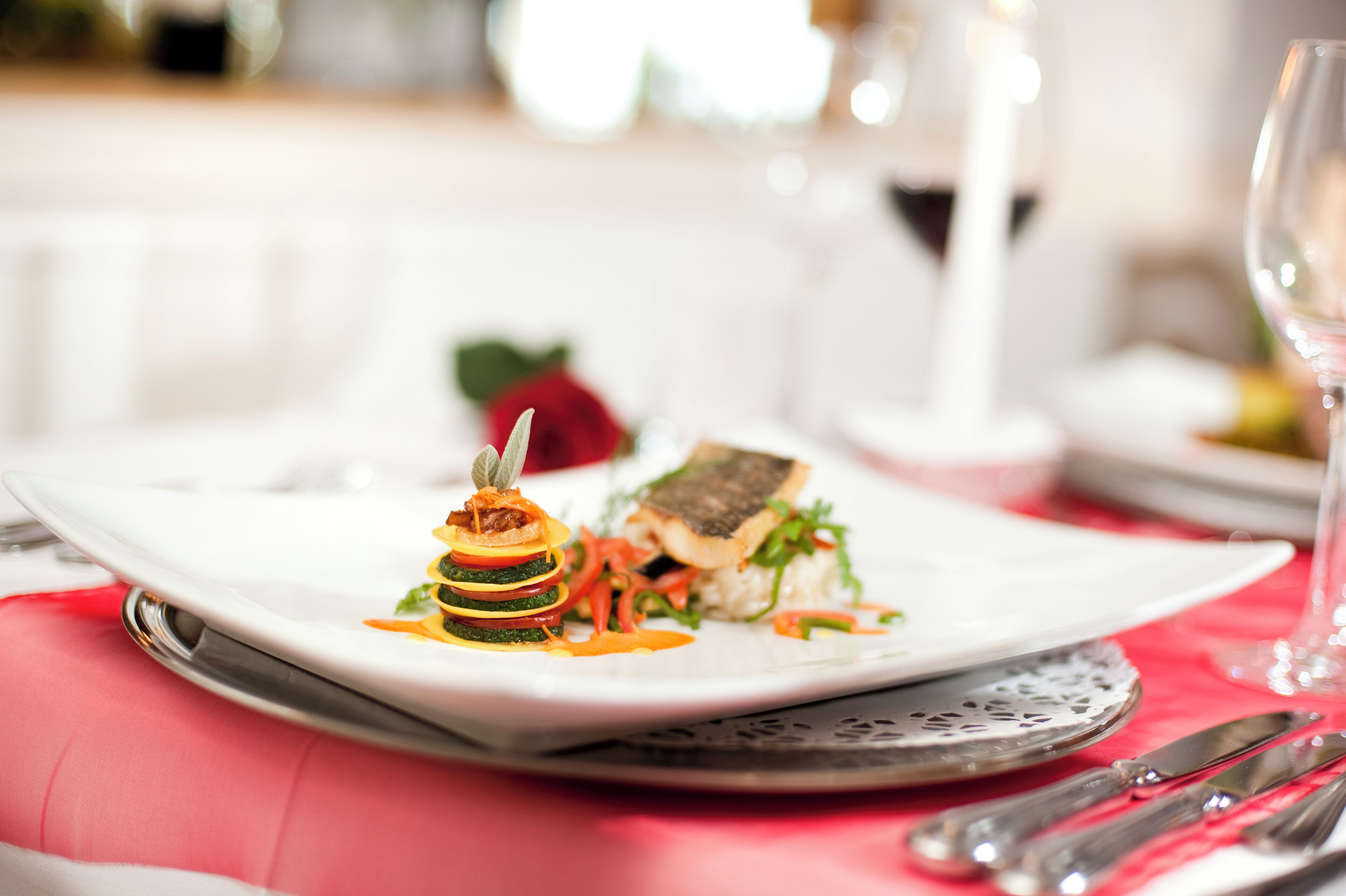 Une assiette blanche contenant un plat de poisson, une rose et un verre de vin, le tout posé sur une table recouverte d’une nappe rouge.