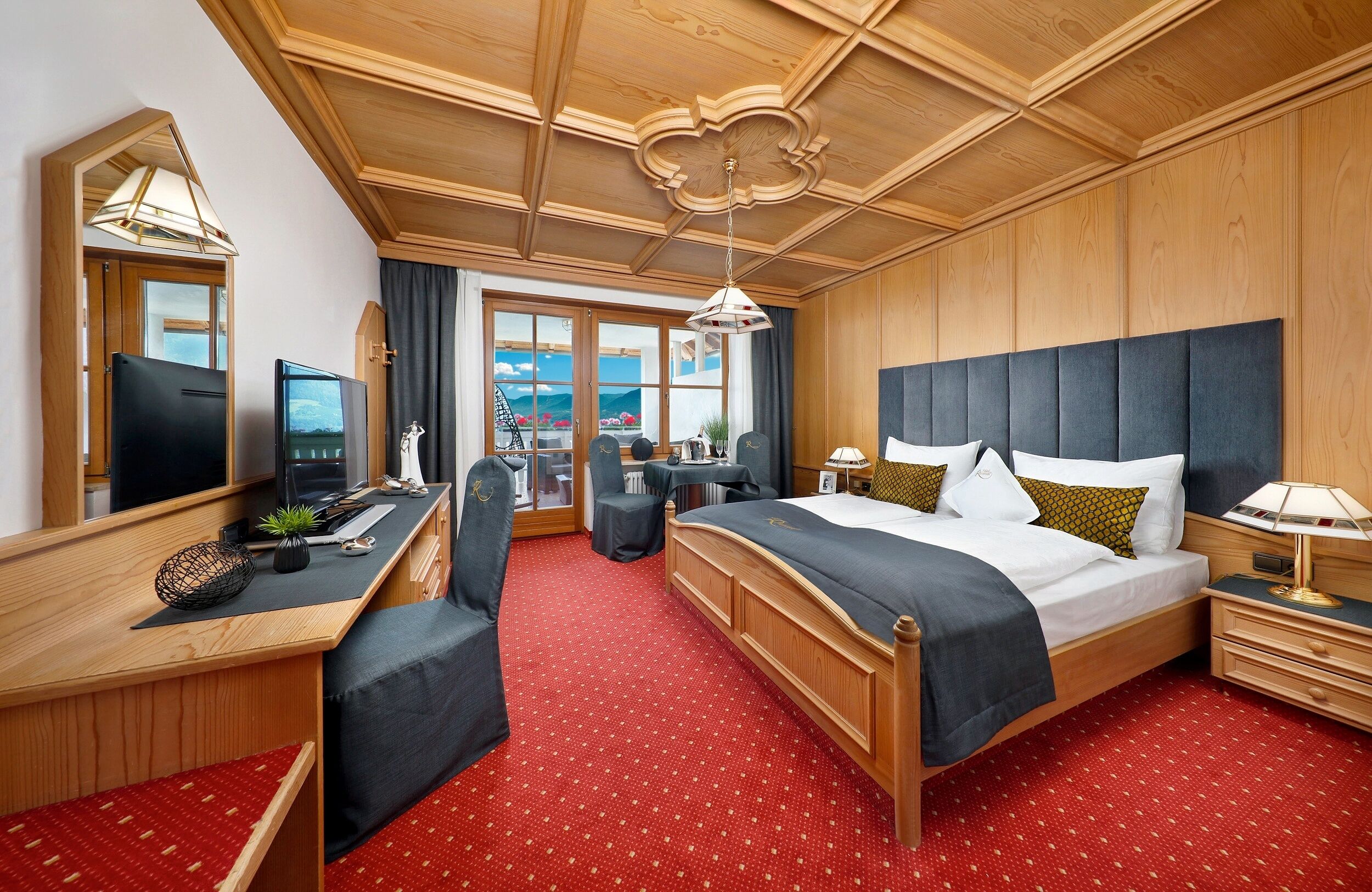 Ein Hotelzimmer mit Holzbalkendecke, einem großen Bett mit dunklem Kopfteil, einem Schreibtisch mit Computer, einem Sessel und Blick ins Freie durch die Fenster.
