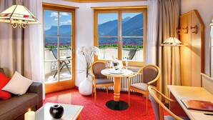 Quarto de hotel com carpete vermelho, sofá com almofadas vermelhas e brancas, mesa de jantar com cadeiras, vaso com flores e vista para montanhas por grandes janelas.