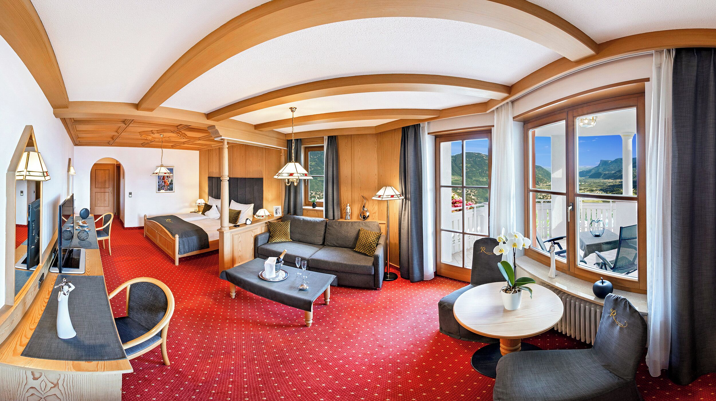 A hotel room with a large bed, a sofa, a dining table, and a balcony with a view of the mountains.