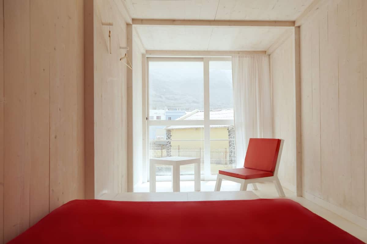 Une pièce avec un lit rouge, une chaise rouge et une vue sur les immeubles, visible à travers une grande fenêtre.