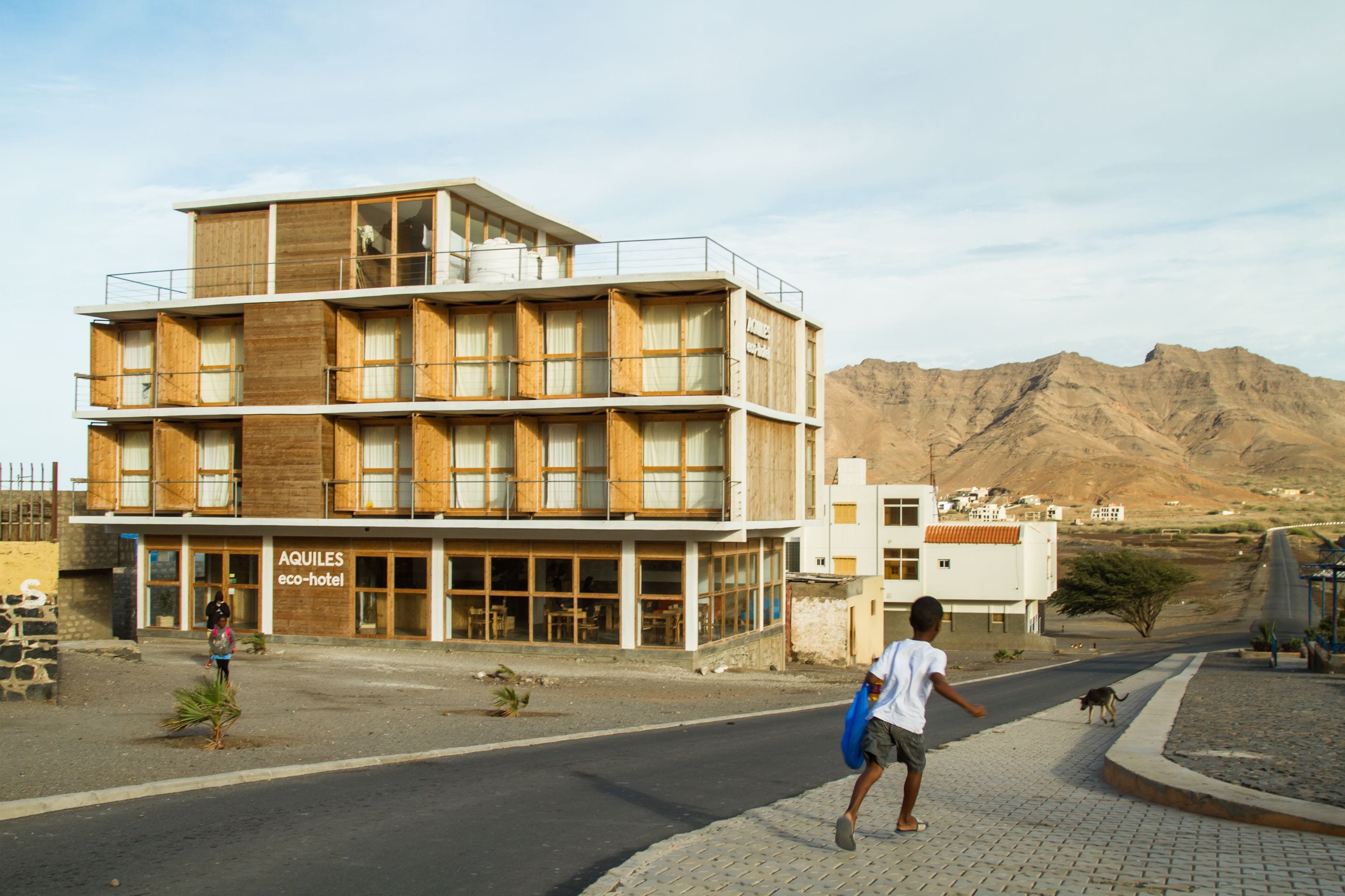 Un immeuble à plusieurs étages doté d’une façade en bois, portant l’inscription « AQUILES, hôtel écologique », et agrémenté de balcons et de grandes fenêtres.