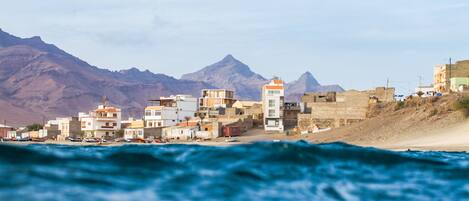 A coastal town with buildings along the shore, a lighthouse, and mountains in the background.