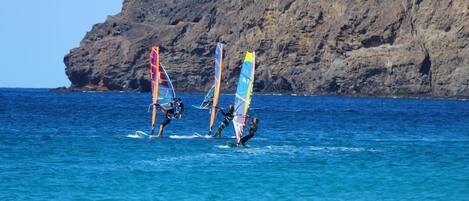 Three windsurfers on a clear blue sea with a rocky cliff and a lighthouse in the background.