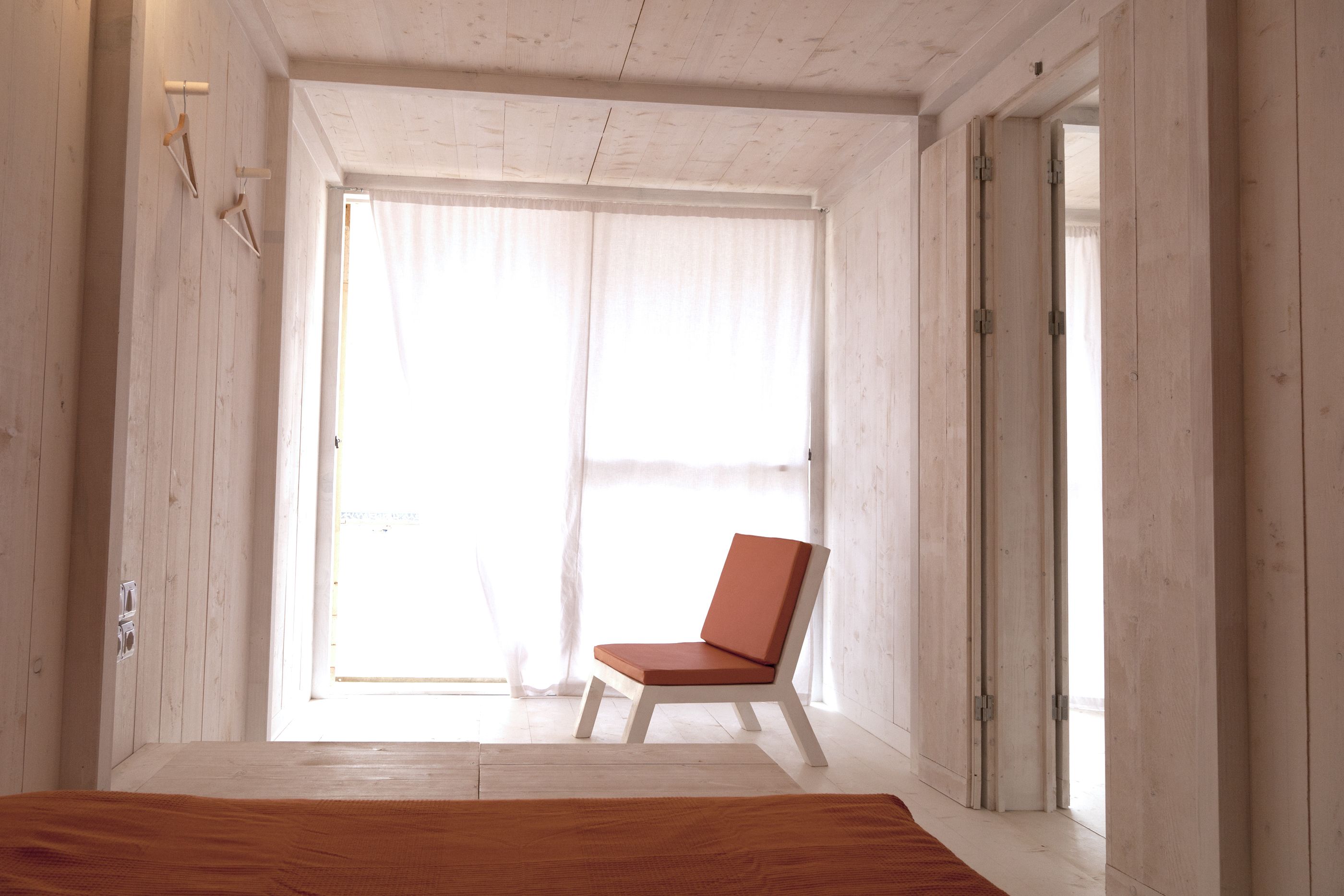 A room with a wooden floor, a white chair, and a large window with sheer curtains.