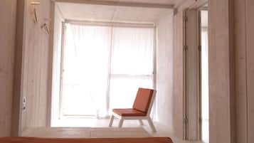 A room with a wooden floor, a white chair, and a large window with sheer curtains.