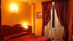 Ein Hotelzimmer mit einem Bett, einem Nachttisch, einer Wandlampe, einem gerahmten Bild und einem Fenster mit Vorhängen.