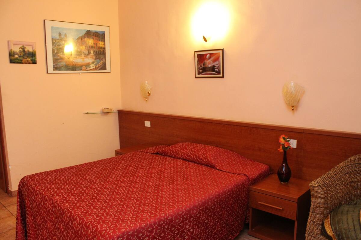 Habitación de hotel con una cama, una mesita de noche, un jarrón con flores y dos lámparas fijadas a la pared.