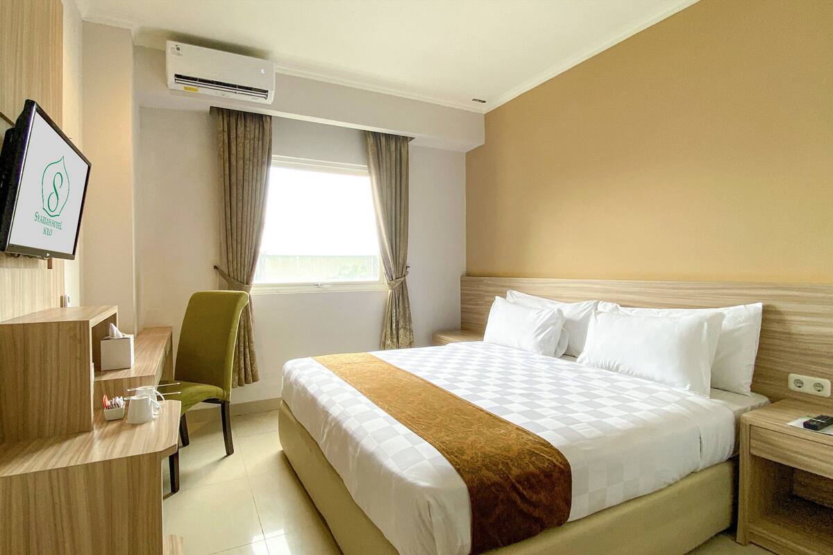 A hotel room with a bed, a desk, a chair, an air conditioning unit, and a window with curtains.