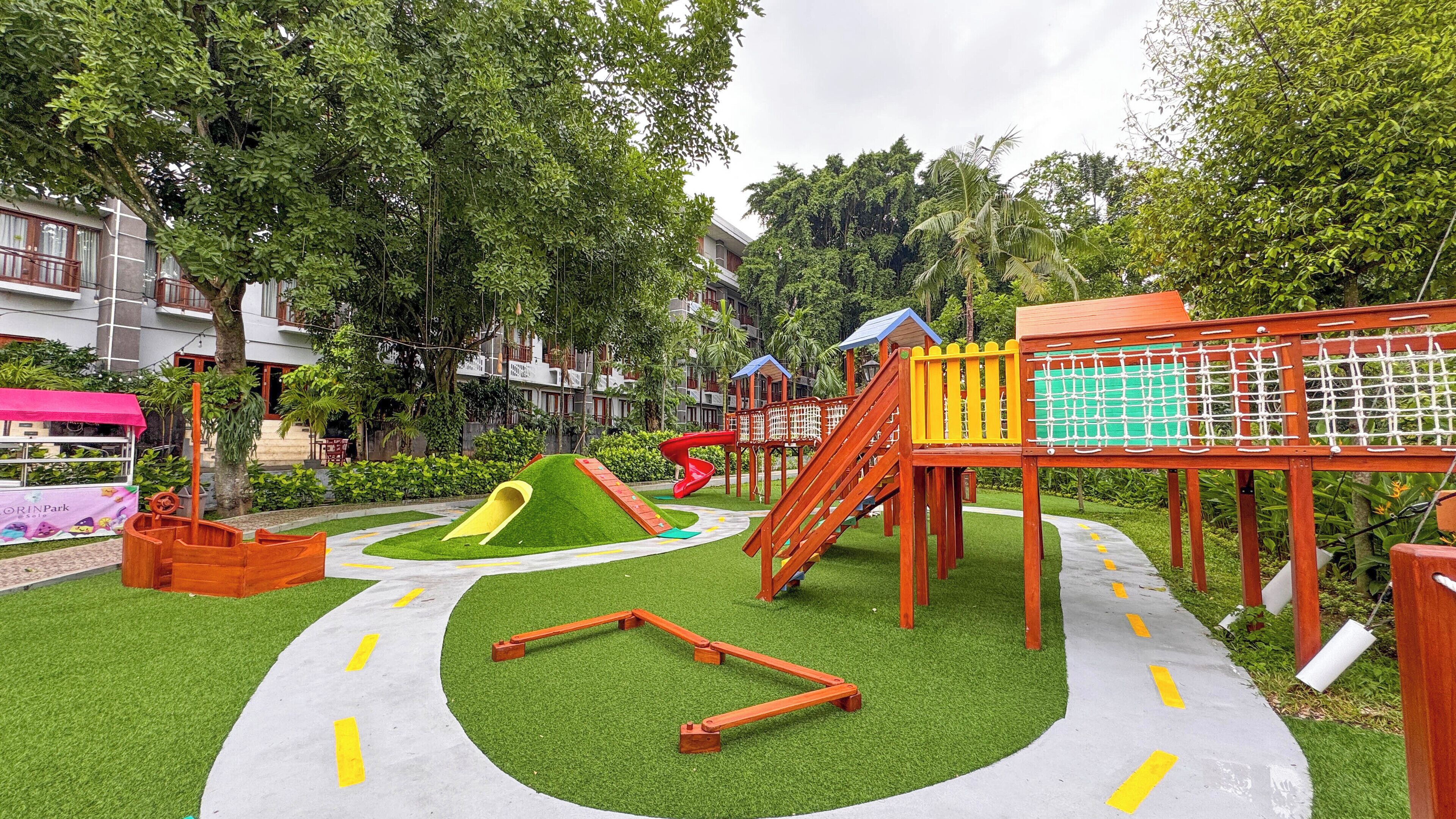 A playground with slides, climbing structures, and a netted area, surrounded by greenery and buildings.