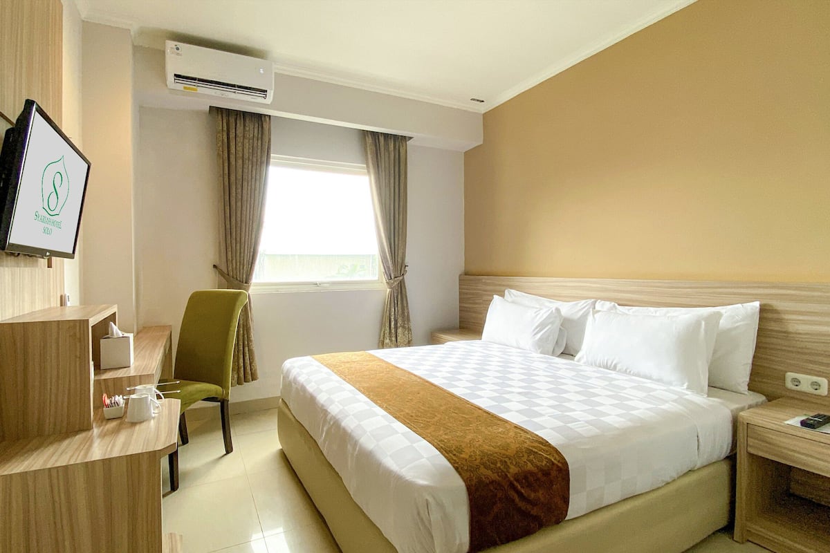A hotel room with a bed, a desk, a chair, an air conditioning unit, and a window with curtains.