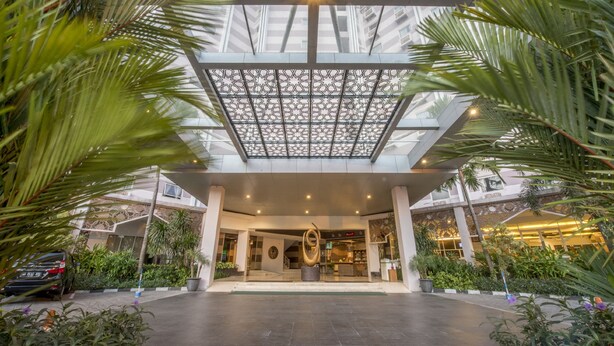 A hotel entrance with a modern glass ceiling, a central sculpture, and surrounding greenery. - Lorin Syariah Solo Hotel (Colomadu)