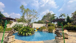 A swimming pool with a slide, surrounded by greenery and stone statues. - Lorin Syariah Solo Hotel (Colomadu)