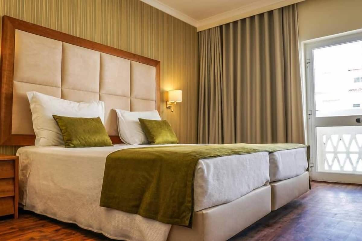 Une chambre d’hôtel avec un grand lit, deux lampes de chevet, une table de nuit en bois et une vue sur l’extérieur par une porte munie de rideaux.