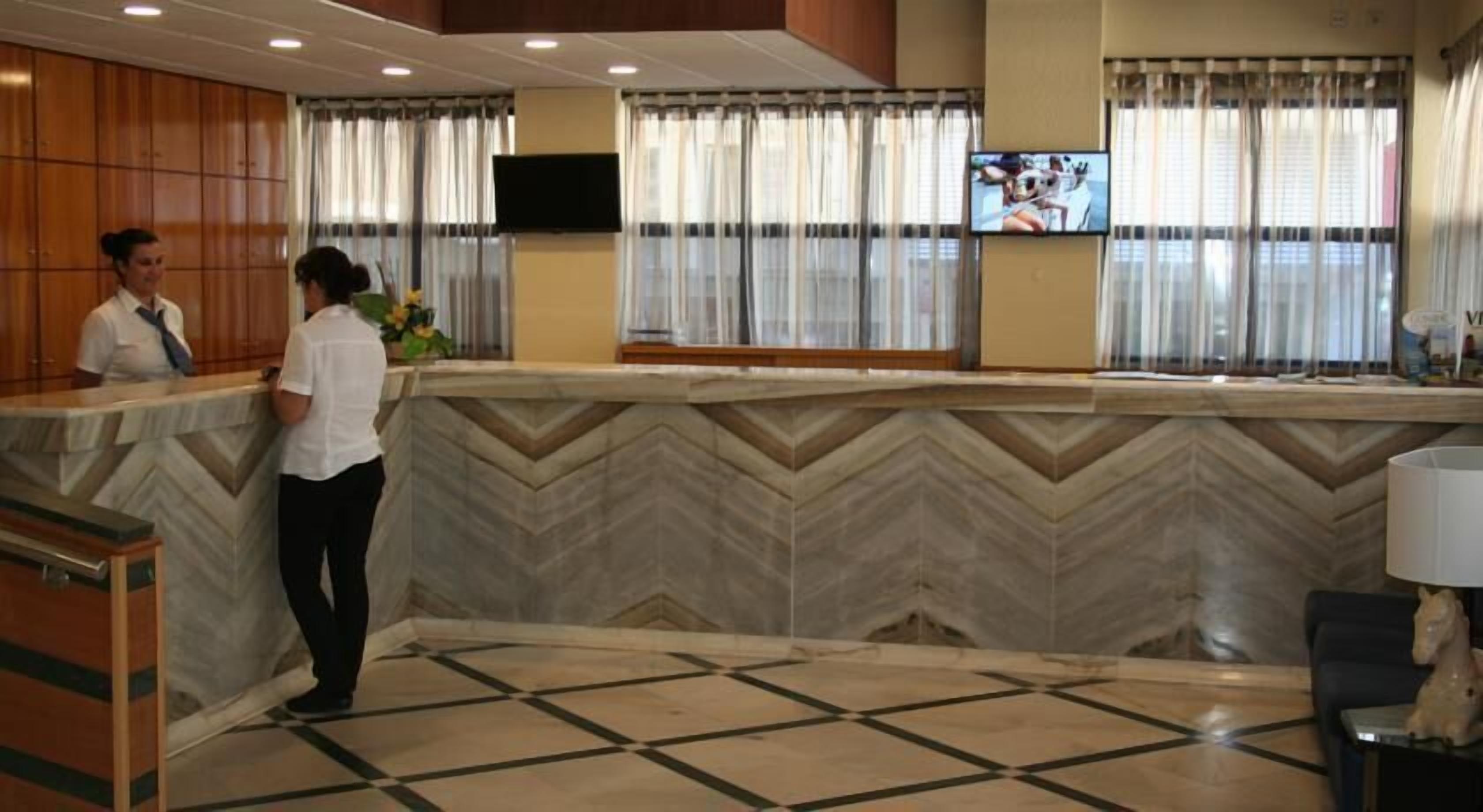 Un área de recepción con una pared de diseño en zigzag, un mostrador de recepción, un televisor de pantalla plana montado en la pared y una ventana con cortinas.