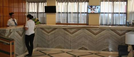 A reception area with a chevron-patterned wall, a reception desk, a flat-screen TV mounted on the wall, and a window with curtains.