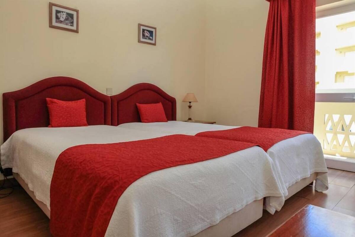 Un lit double avec une literie rouge et blanche, deux tableaux encadrés au mur, une table de chevet avec une lampe et un rideau rouge.
