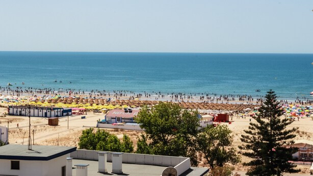 A beach with numerous umbrellas and people. There are buildings with signs for a restaurant and a grill. - Baía Beach Hotel (Vila Real Santo Antonio)
