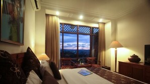 Habitación de hotel con una cama grande, un escritorio, una silla, una lámpara y vistas a la ciudad a través de una ventana.
