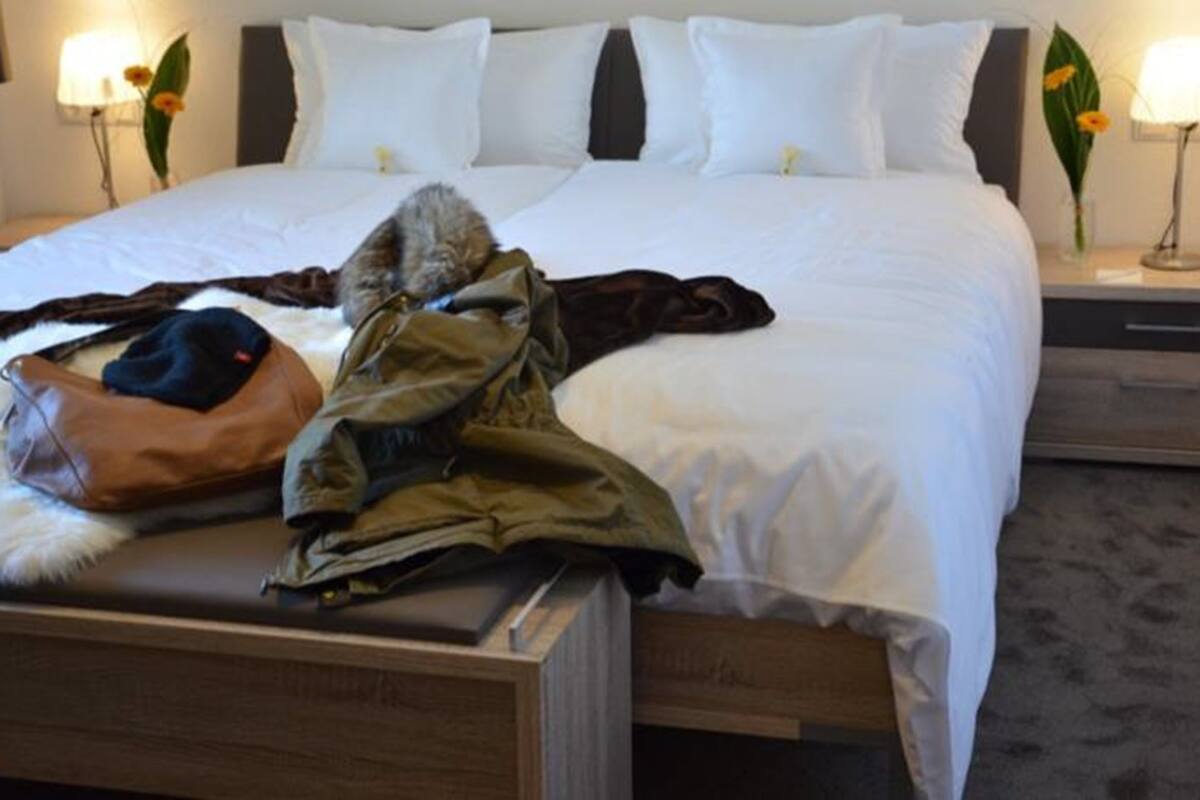 Uma cama bem arrumada com lençóis brancos, uma mala de couro marrom e um casaco verde sobre a cama.