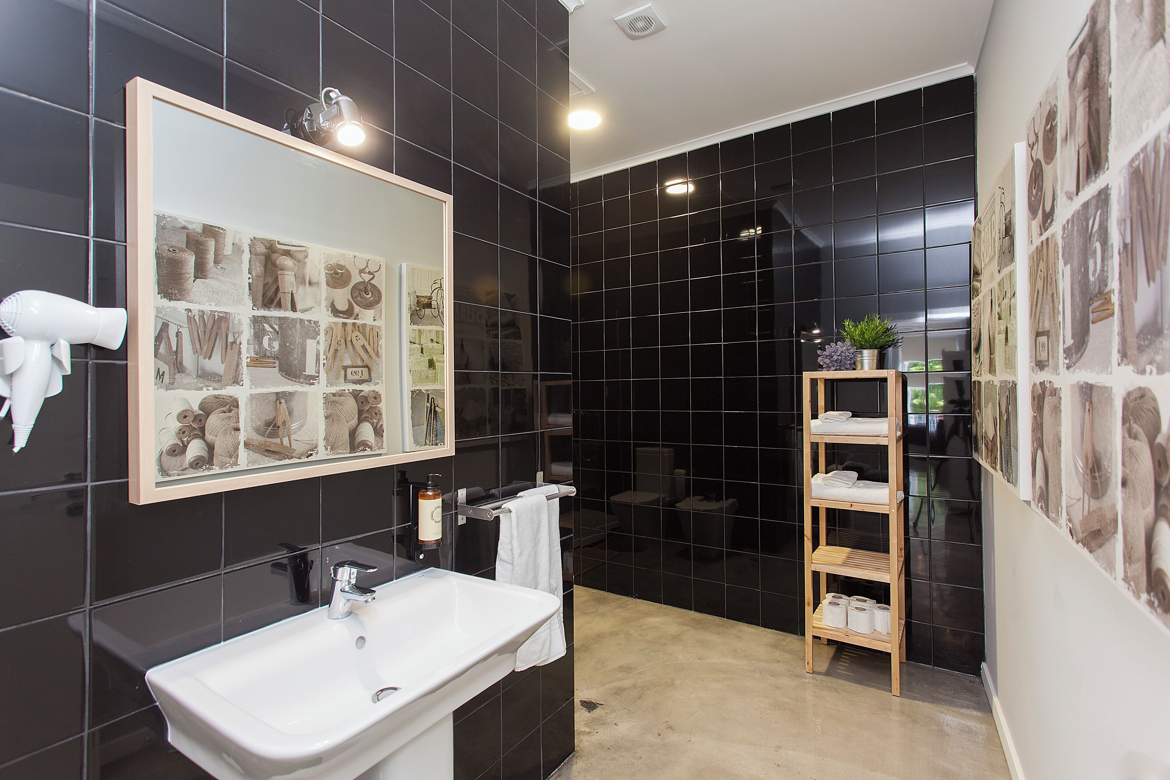 Un baño moderno con un espejo grande, azulejos negros, un lavamanos blanco y un estante de madera con toallas.