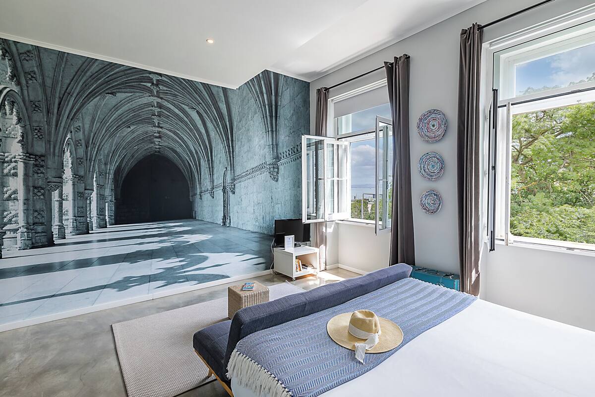 A room with a large mural of a cathedral's interior, a bed with a blue and white striped bedspread, a wicker side table, and a view of greenery through a window.