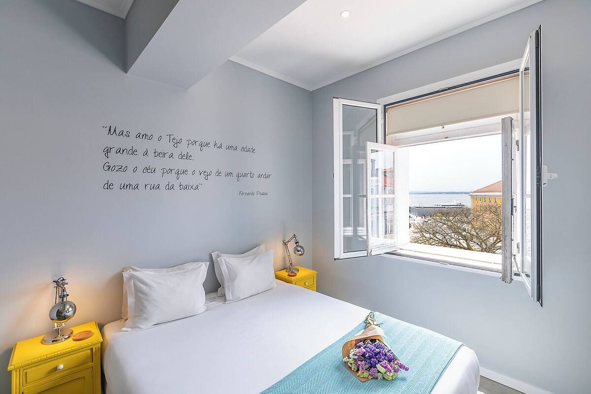 A bedroom with a bed, a nightstand, a lamp, and a window with an open view.