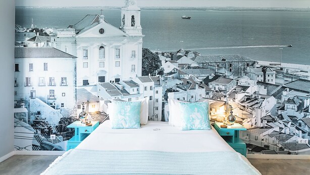 A bedroom with a mural of a coastal town, a bed, and bedside tables with lamps. - Emporium Lisbon Suites (Lisbon)