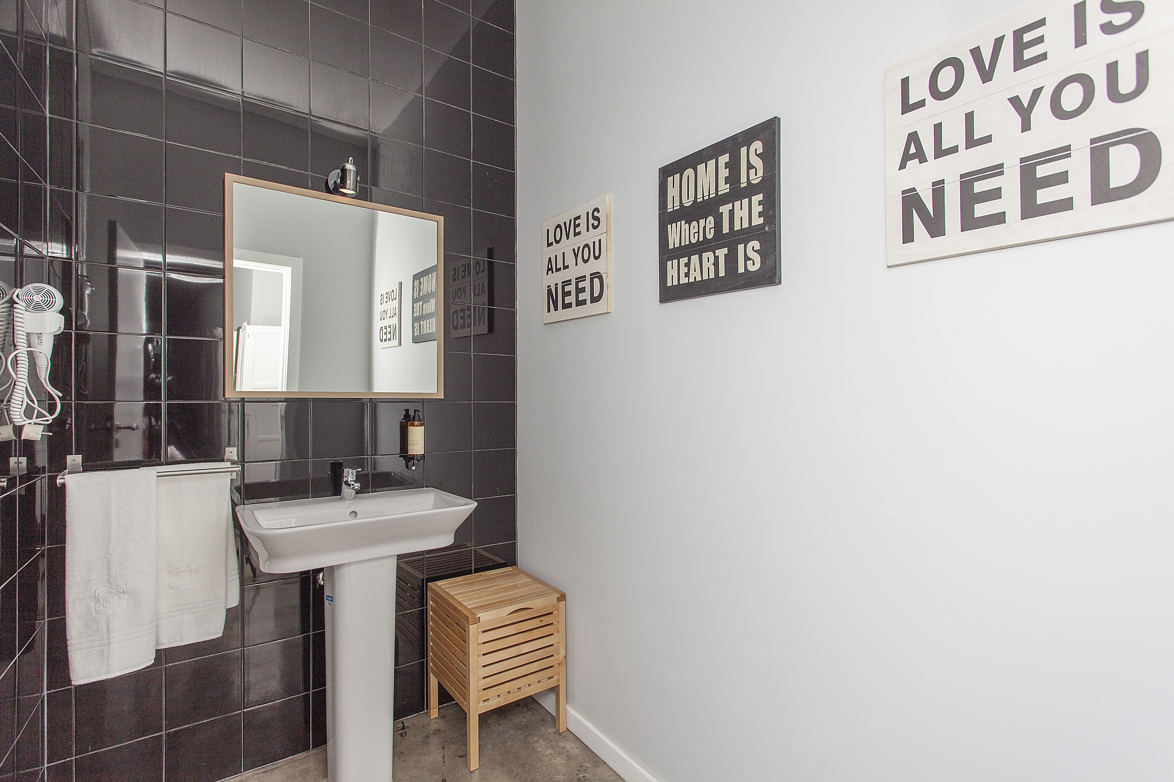 Un baño moderno con lavamanos blanco, baldosas oscuras, un taburete de madera y arte decorativo en la pared.
