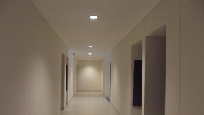 A narrow, well-lit hallway with white walls and tiled flooring, leading to a darkened room at the end. - L Bajo Hotel (Labuan Bajo)