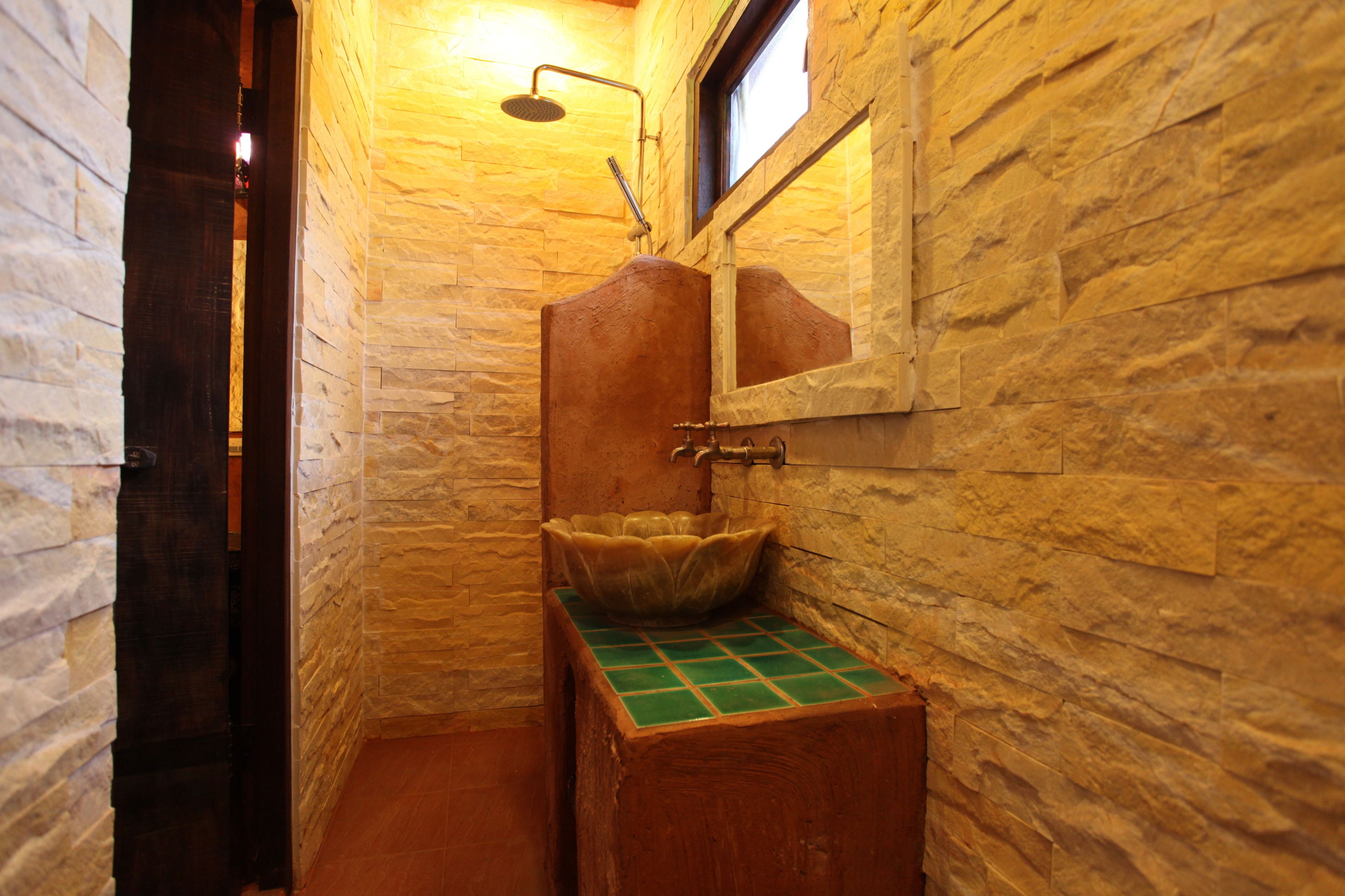 Un baño con pared de piedra, un lavabo de madera y encimera verde de azulejos, y una ducha con cabezal de ducha manual.