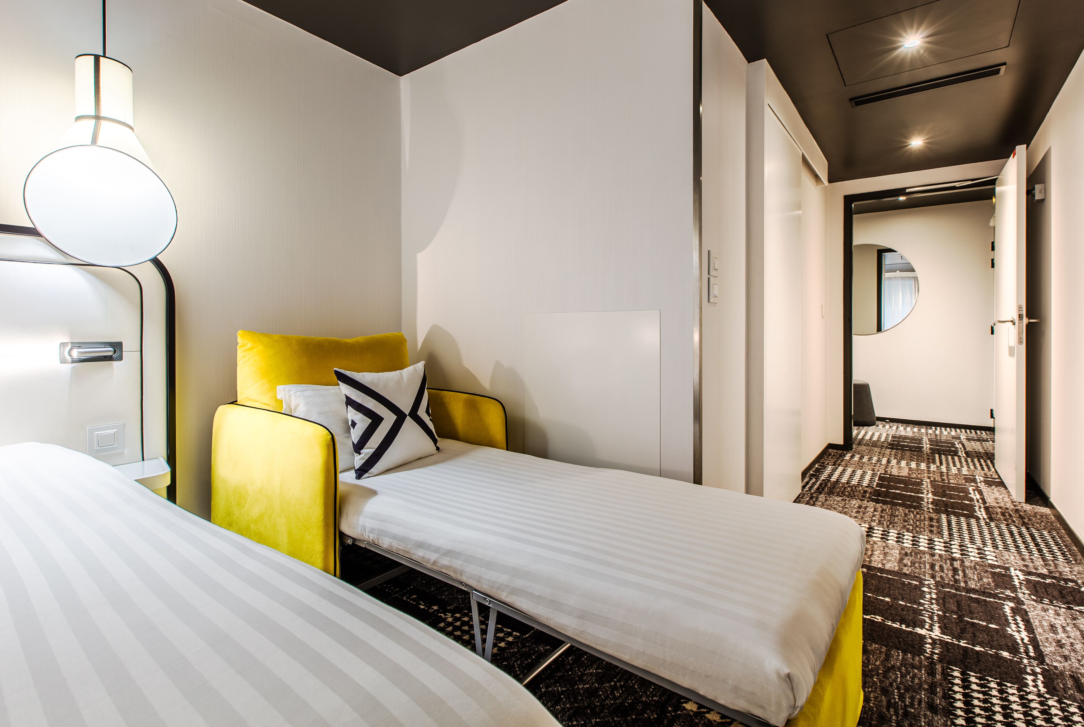 Habitación de hotel con dos camas individuales, un sillón amarillo, una mesita de noche, una lámpara y una alfombra estampada.