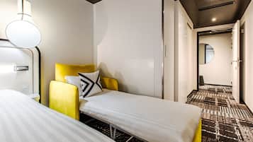 Habitación de hotel con dos camas individuales, un sillón amarillo, una mesita de noche, una lámpara y una alfombra estampada.