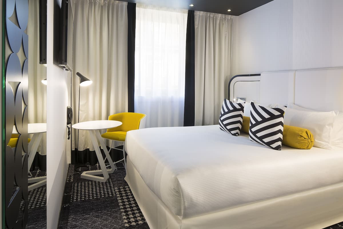 A hotel room with a large bed, a small round table, a yellow chair, and a mirror.