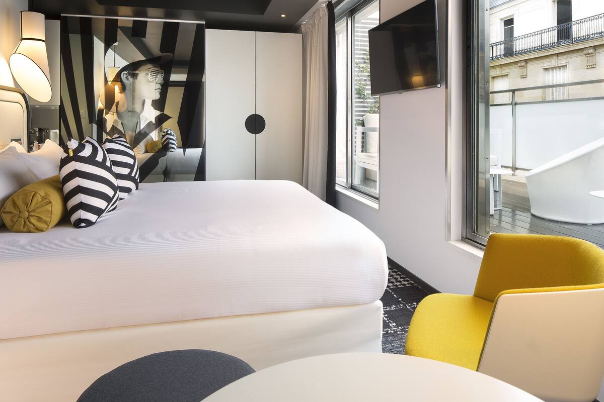 A modern hotel room with a large bed, a yellow armchair, and a large window offering a view of the city.