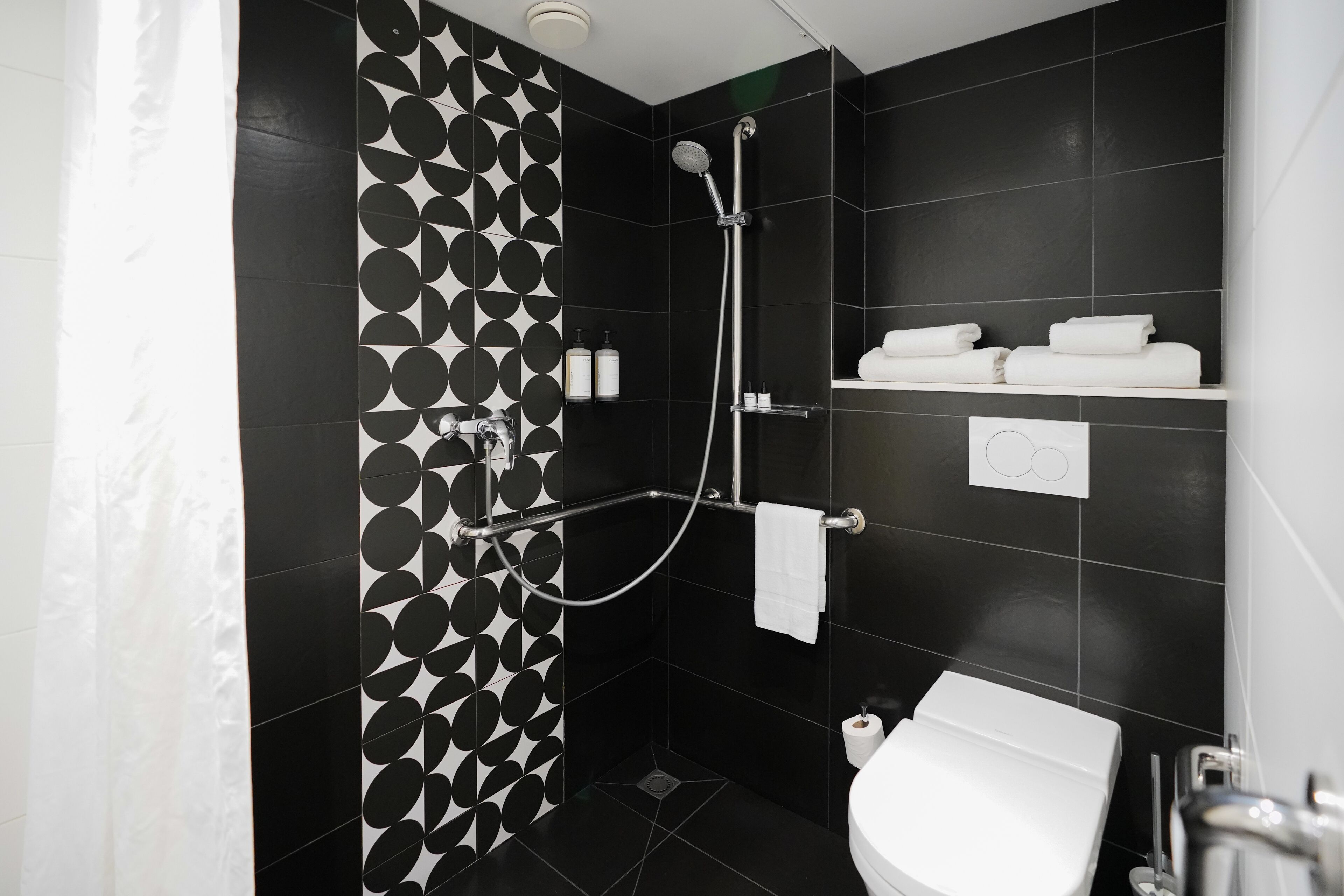 Casa de banho moderna com azulejos preto e branco, chuveiro, sanita e um prateleira com toalhas.
