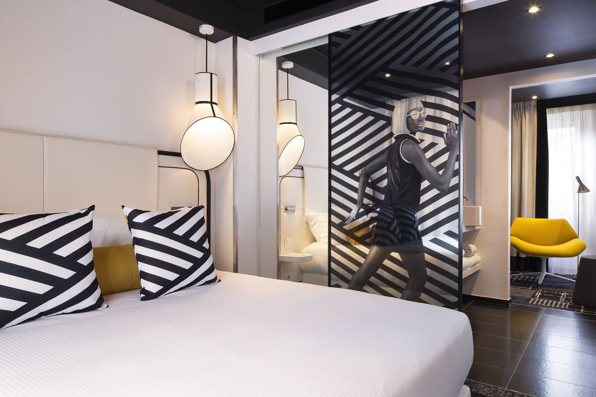 A modern hotel room with a large bed, a mirror, a small bedside table, and a yellow chair.