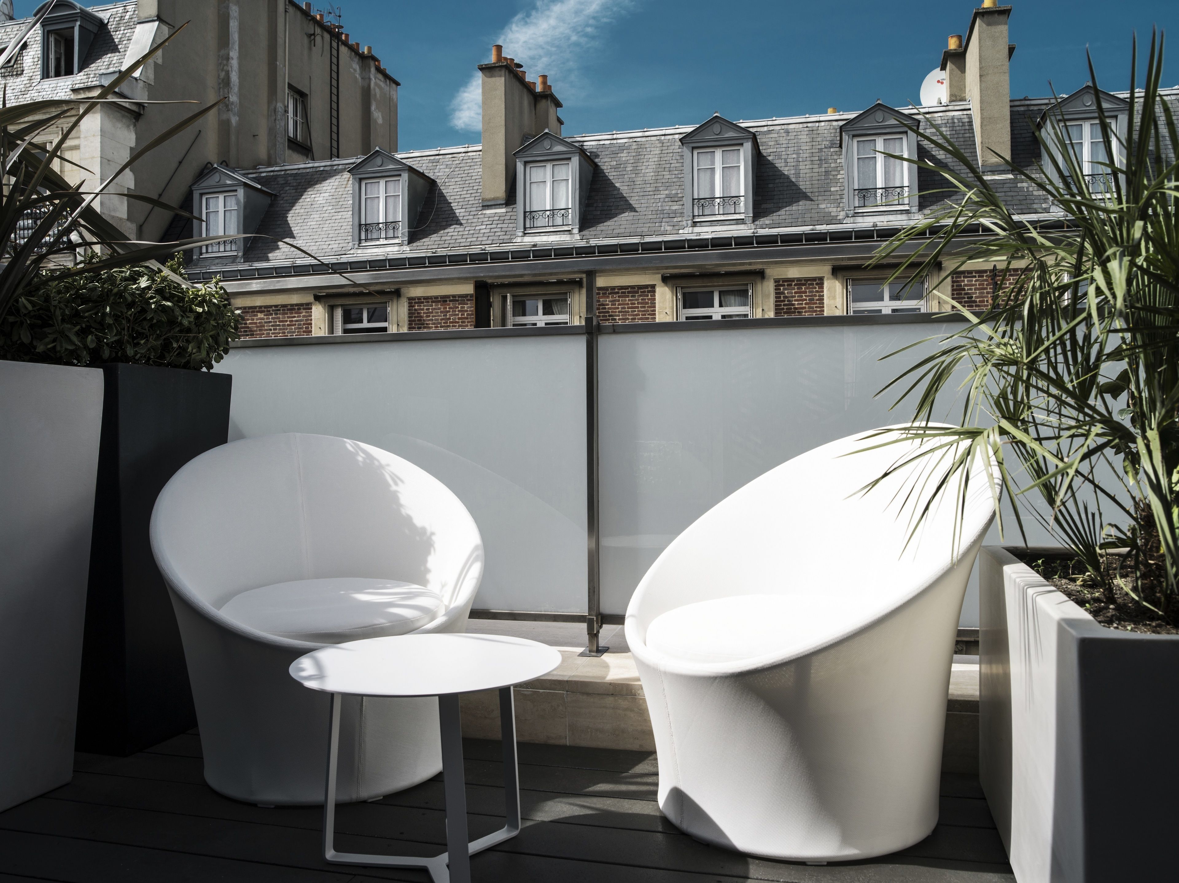 A rooftop terrace with two white lounge chairs and a small table, surrounded by potted plants.