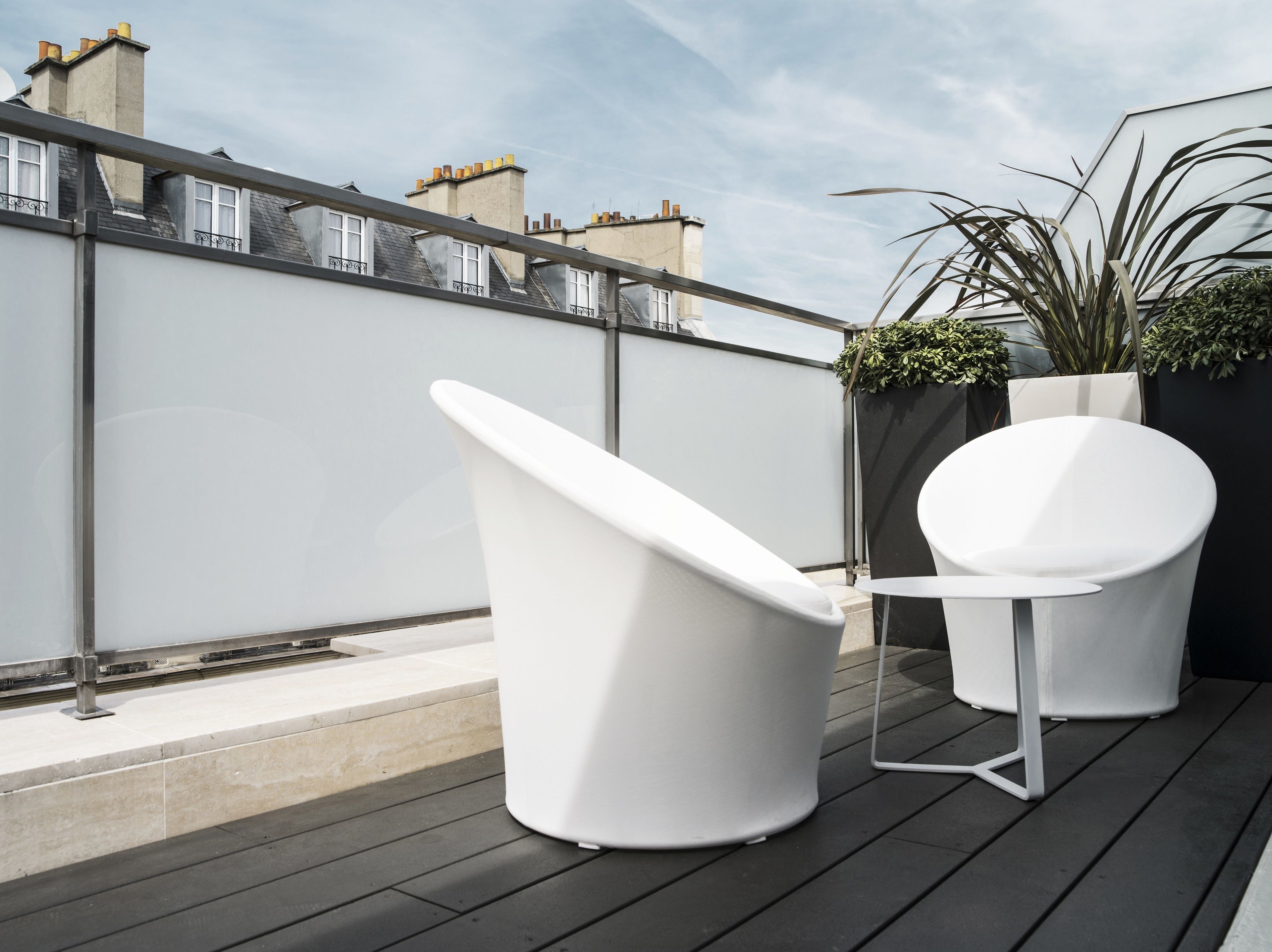 A rooftop terrace with two white chairs and a small table, surrounded by a low wall and potted plants.