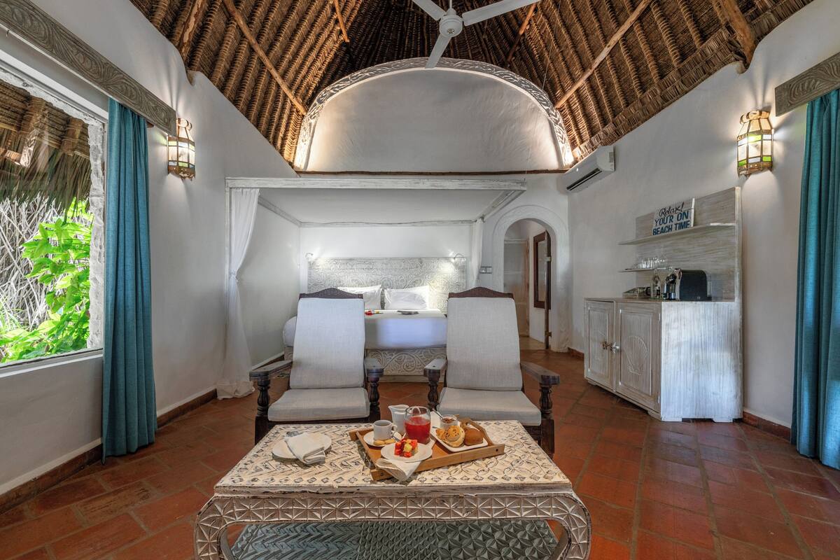 A room with a bed, two chairs, a dining table with breakfast, and a kitchen area.