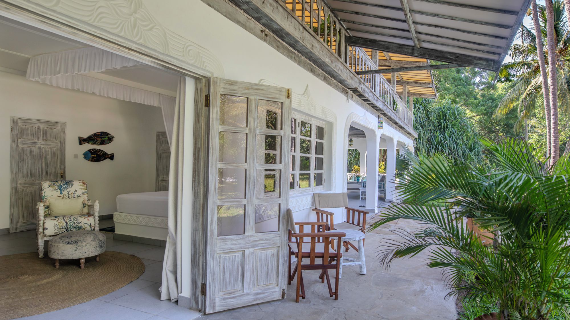 A room with a bed, a chair, and a table. There is a balcony with a railing and a view of greenery.