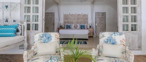 A room with a bed, two chairs, a small table with a plant, and a view of another room through an open door.