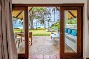 Vista a través de una puerta de madera a un patio con suelo de piedra, mobiliario exterior y vista a la playa.